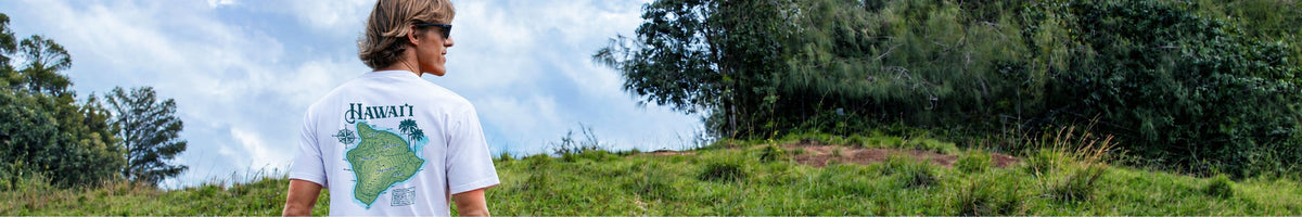 A man standing outdoors on a grassy hill wearing a white t-shirt with a green map graphic and the word 'Hawaii' printed on the back, looking up at a cloudy sky with trees in the background.