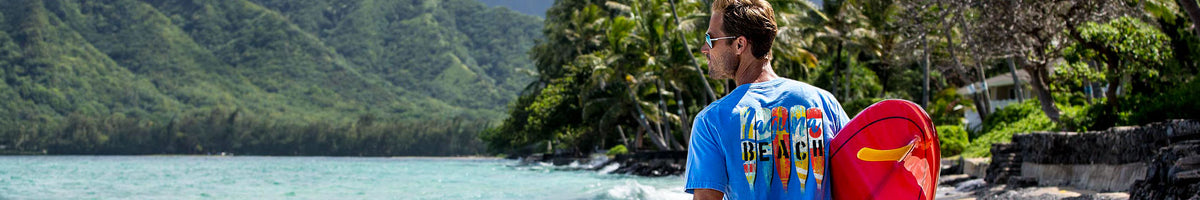 A man wearing a blue Hawaii surf shirt stands on a beach holding a red and yellow surfboard, with a lush green tropical coastline and ocean in the background.