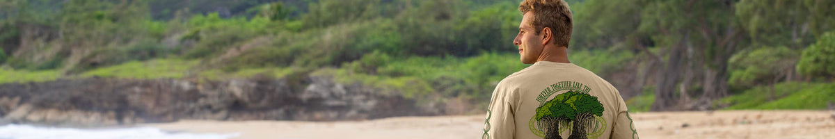 A man standing on a sandy beach facing a lush green forest and rocky coastline, wearing a beige t-shirt with a large green graphic print on the back.