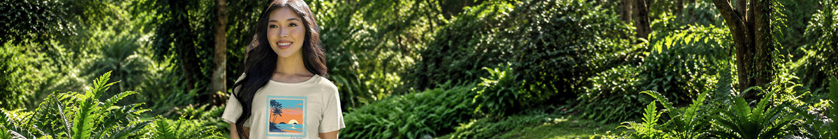 A woman wearing a beige graphic t-shirt stands in a lush green forest setting. The t-shirt features a colorful square graphic on the front depicting a beach scene with blue water, white sand, and a yellow sun.