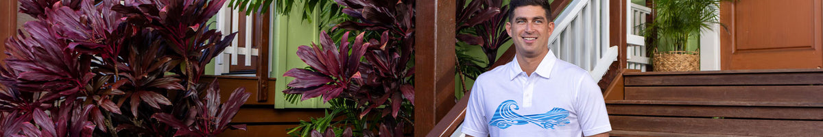 A man wearing a white short-sleeve polo shirt with a blue wave graphic on the front, standing on outdoor wooden steps next to large purple and green foliage.