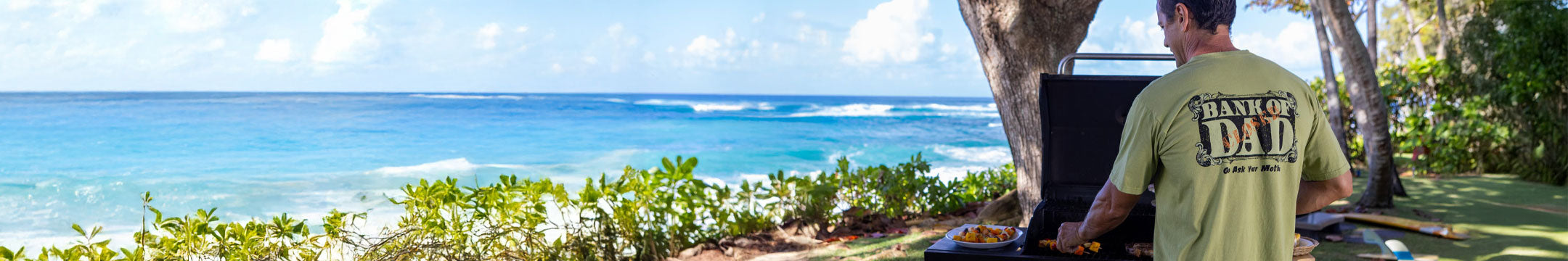 Man in a green shirt with 'Dad' printed on it sitting by a scenic beach.