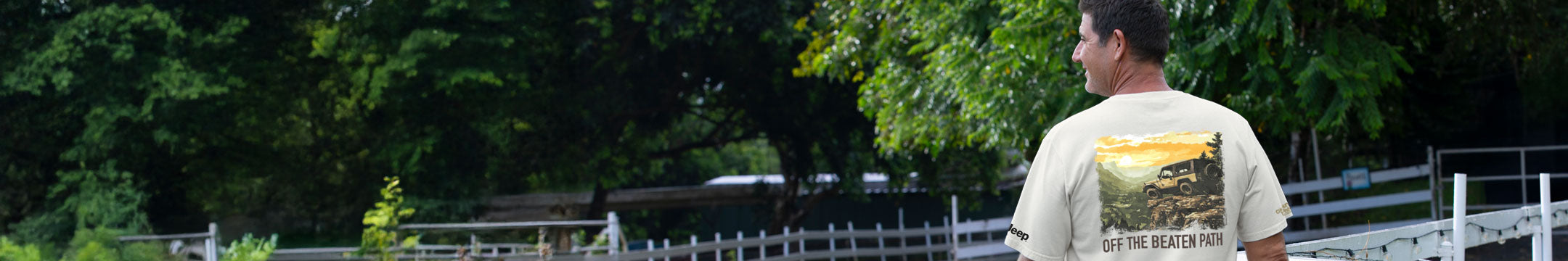 Man wearing a t-shirt with a scenic design, standing outdoors with trees and a fence in the background.