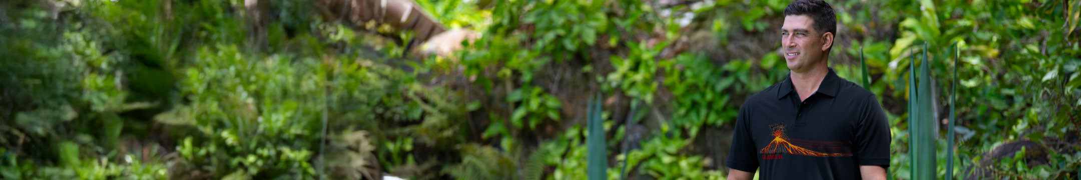 man wearing a black collared shirt standing in front of green foliage.