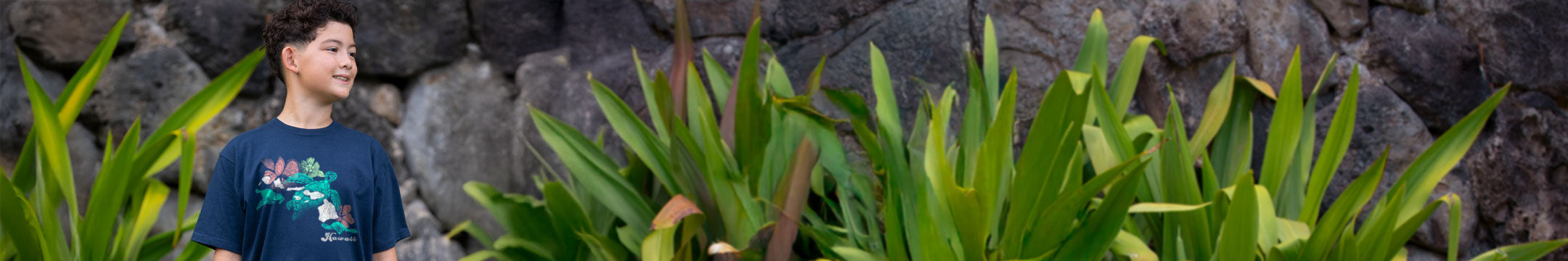 Person wearing a blue shirt with a graphic design, standing in front of a stone wall and green plants.