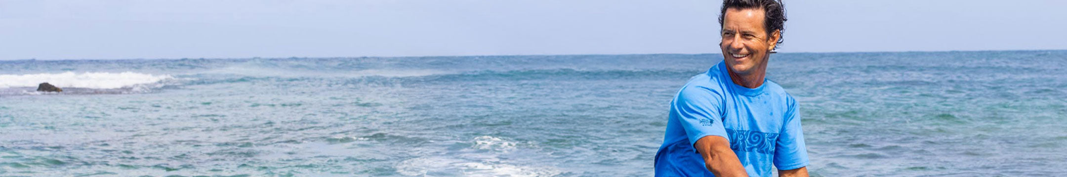 Man in a blue shirt sitting on a rock by the ocean with clear blue water and sky.