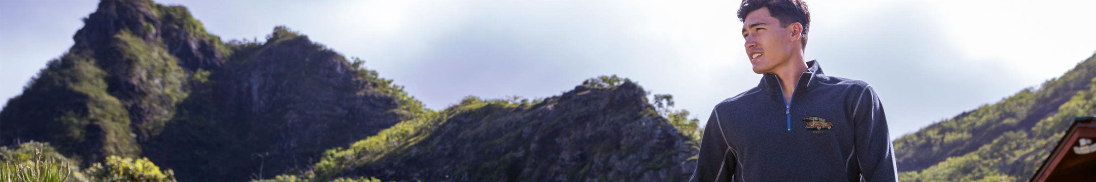 Person wearing a dark jacket with a logo, standing in front of a mountainous landscape.