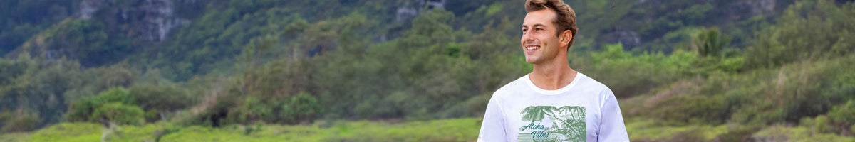 A man wearing a white crewneck t-shirt with a green tropical beach graphic, standing outdoors in front of a lush green mountain landscape.