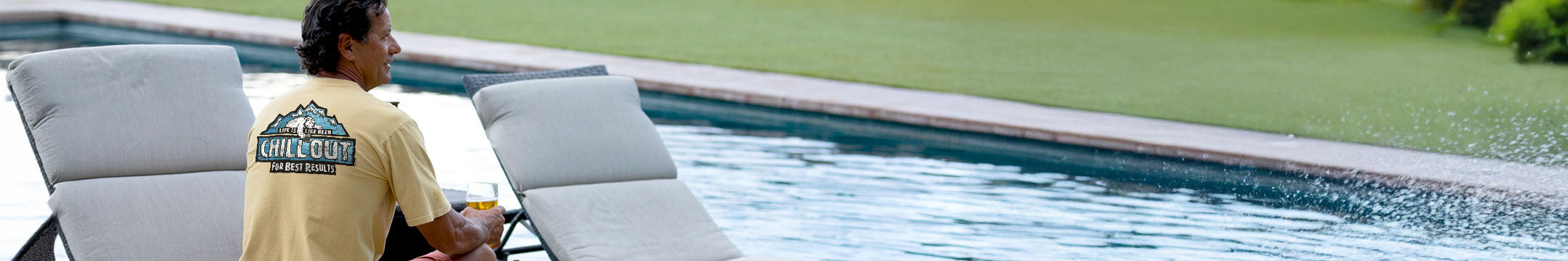 Personwearing a Beer Dyed Crazy Shirt tee sitting by a pool with lounge chairs and a scenic background