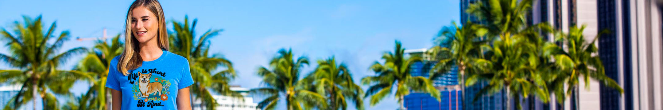 Woman wearing a blue t-shirt with a graphic design, standing in front of palm trees and a building.