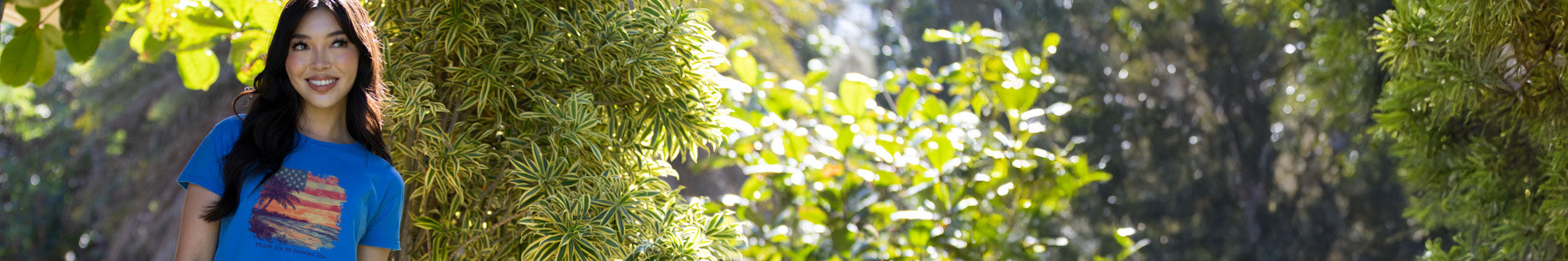 Girl wearing a blue graphic tee with a patriotic print in front of tropical foliage.