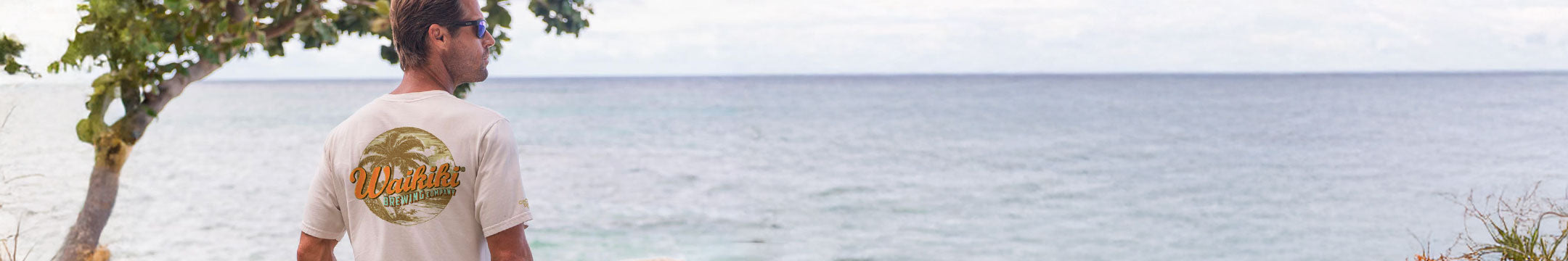 Man wearing a t-shirt with a logo standing on a beach looking at the ocean.