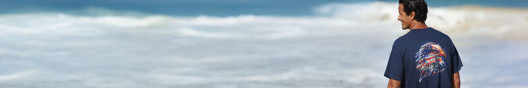 Person wearing a blue t-shirt with a graphic design of an eagle standing on a beach with ocean waves in the background.