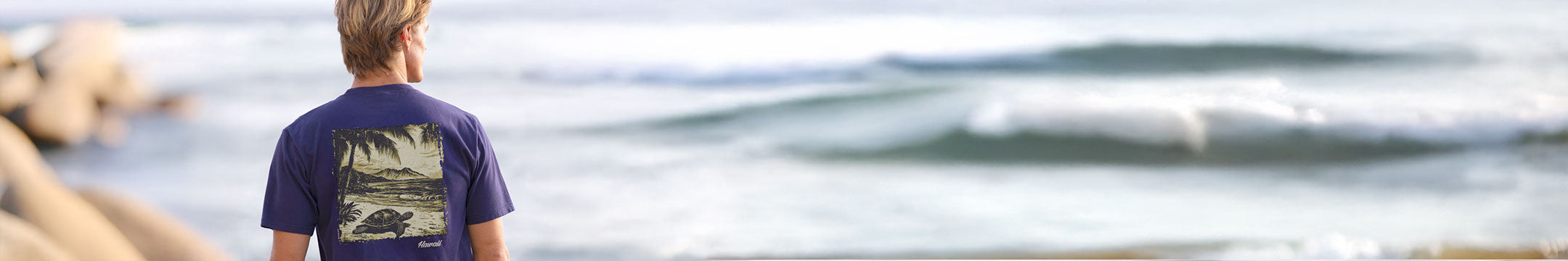 Person wearing a Grape Dyed t-shirt with a graphic design, standing by a body of water.