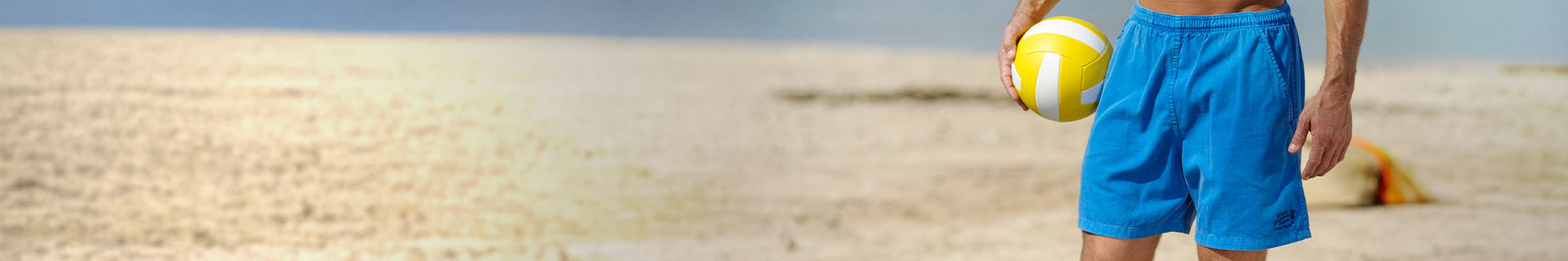 A man wearing blue shorts standing on a sandy beach holding a yellow volleyball. 