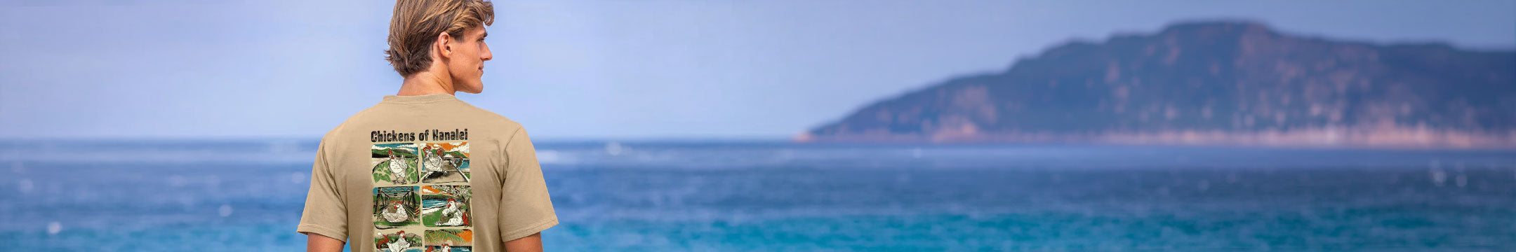 A man stands on a beach facing the ocean, wearing a beige graphic t-shirt with a green and white print on the back. A hazy island is visible in the background under a pale blue sky.