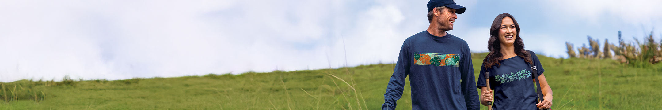 Two people walking outdoors on a grassy hill under a cloudy sky, wearing matching dark blue/gray long-sleeve and short-sleeve shirts with a colorful flower graphic.