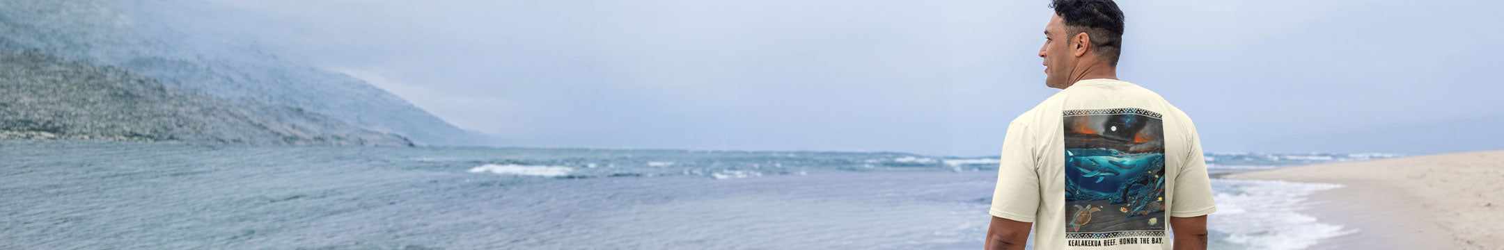 A man standing on a beach wearing a cream-colored t-shirt with a large graphic print on the back, looking out towards the ocean and mountains under a cloudy sky.