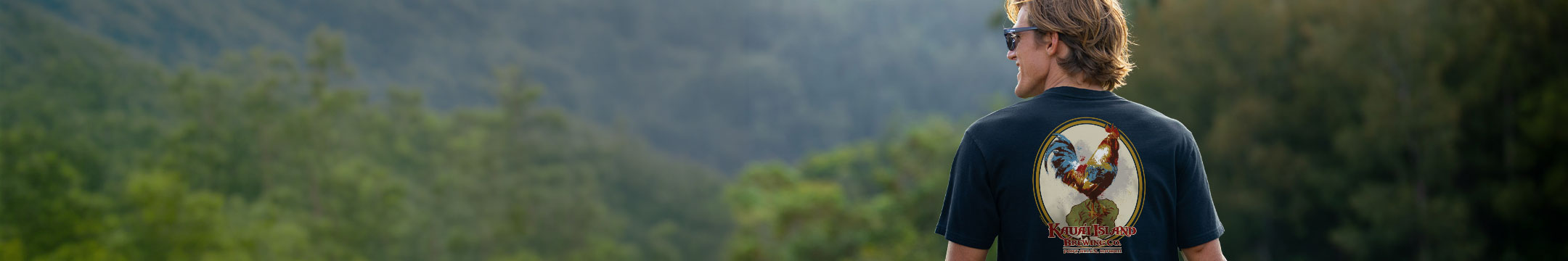 A man viewed from behind wearing a black graphic t-shirt with a large rooster illustration on the back, set against a blurred green forest background.