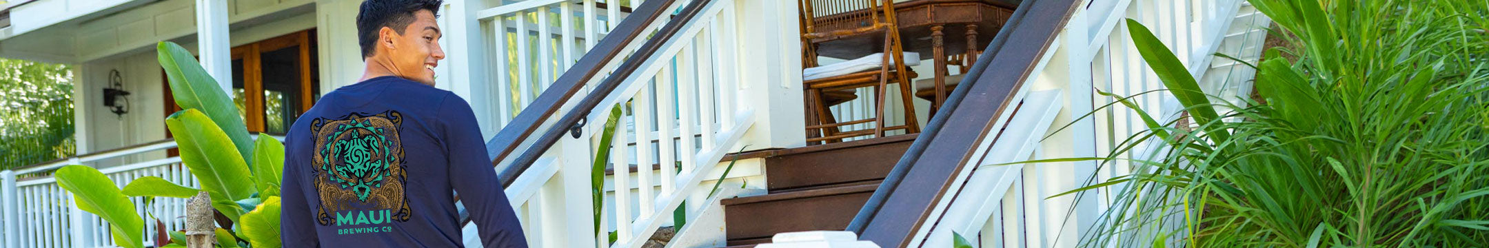 A man wearing a navy blue long-sleeve shirt with a green and brown Maui graphic walks down outdoor wooden stairs next to a white house with tropical plants.