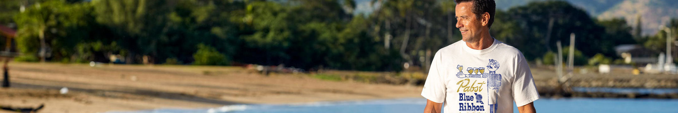 A man wearing a white graphic t-shirt stands on a sandy beach with water and trees in the background.