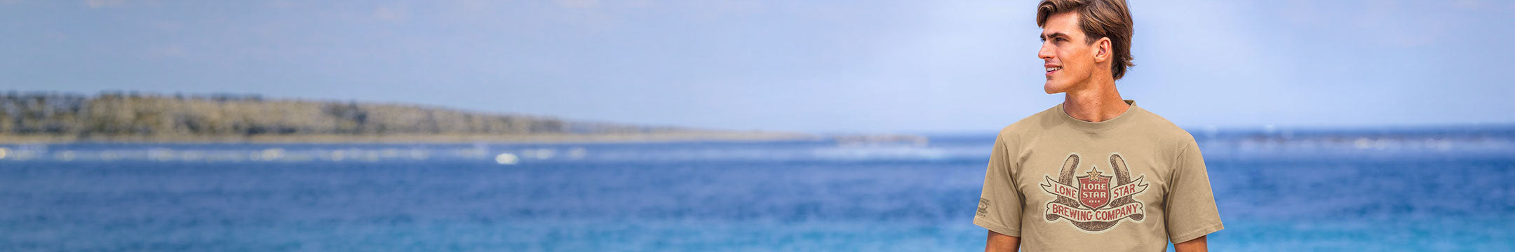 A man wearing a beige graphic t-shirt stands on a beach with blue ocean water and a distant green shoreline in the background.