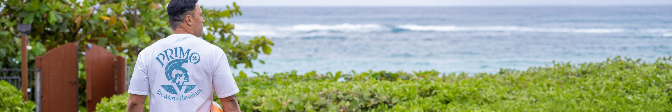 A man standing outdoors by the ocean, seen from behind, wearing a white t-shirt with a blue graphic print that reads 'PRIMO' and shows the back of the shirt.