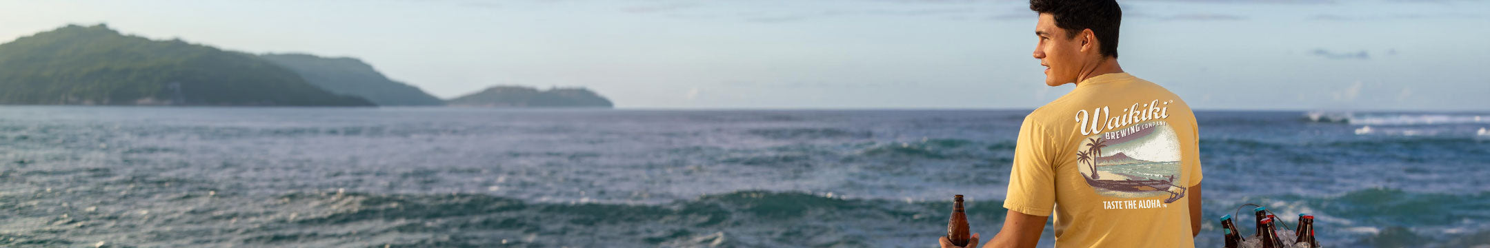 A man wearing a yellow graphic t-shirt stands on a beach overlooking the ocean, with the word 'Waikiki' printed on the back of the shirt.