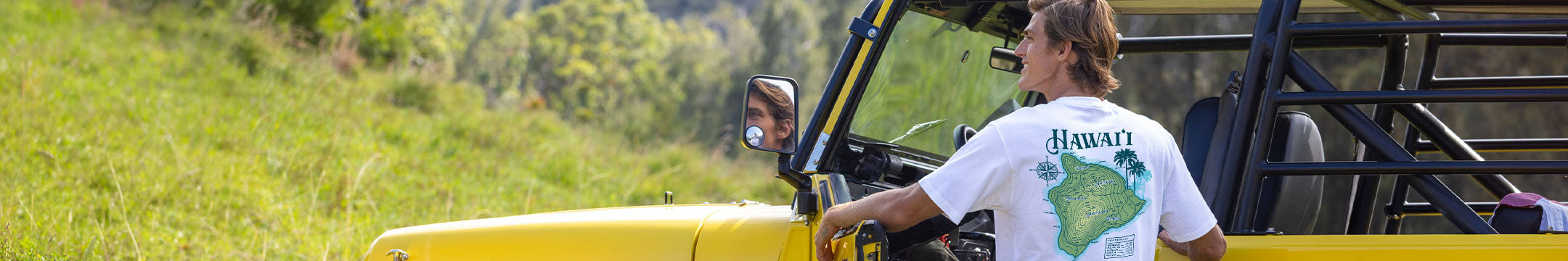 A man wearing a white t-shirt with a green map graphic and the word 'HAWAII' on the back, standing next to a yellow off-road vehicle in a grassy outdoor setting.
