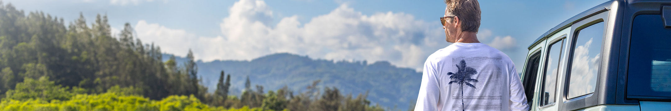 A man wearing a white t-shirt with a black palm tree graphic on the back stands next to a blue vehicle, looking out at a scenic mountain and forest background.