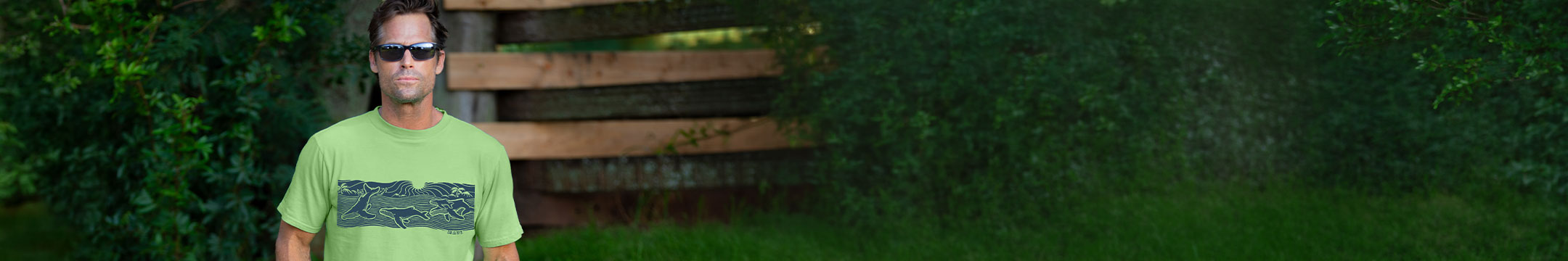 A man wearing a light green t-shirt with a dark graphic print stands outdoors in front of a wooden fence and green foliage.