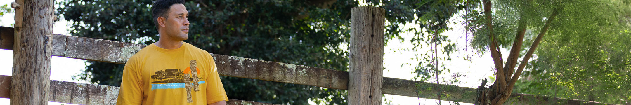 A man wearing a yellow graphic t-shirt with tiki statues, standing outdoors in front of a rustic wooden fence and green trees.
