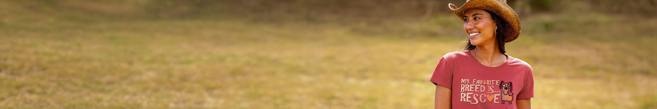 A woman wearing a brown cowboy hat and a light red graphic t-shirt stands in a grassy field. The t-shirt features a graphic with the words 'MY FAVORITE BREED IS RESCUE' in white text.