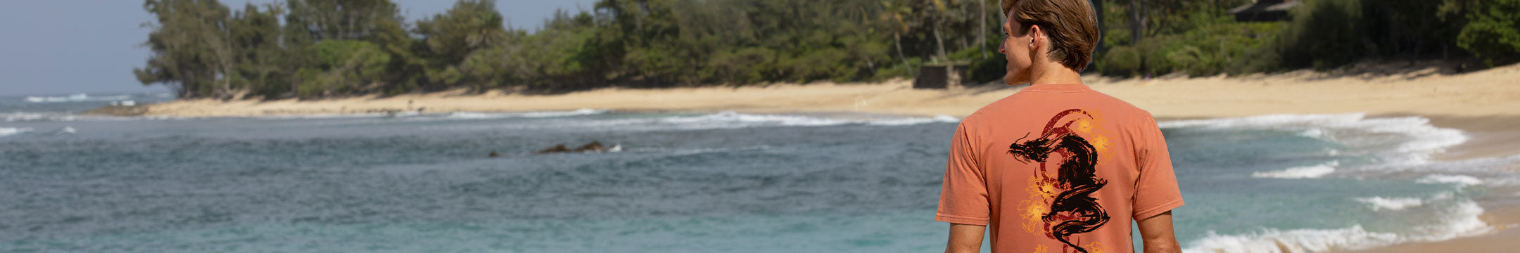 A man wearing a coral orange t-shirt with a black dragon graphic on the back, standing on a tropical beach with turquoise water and green trees in the background.