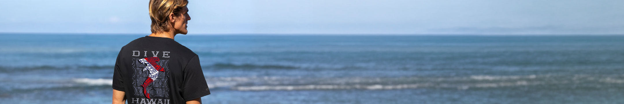 A man wearing a black graphic t-shirt with a tribal shark stands with his back to the camera, looking out towards the ocean under a clear blue sky.