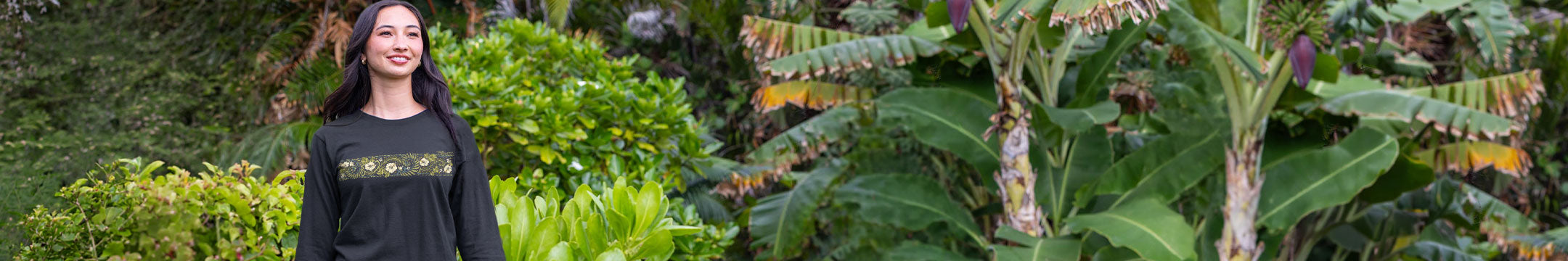 A woman with long dark hair stands outdoors in front of lush green tropical plants, wearing a black long-sleeve top with yellow flower band on the chest.