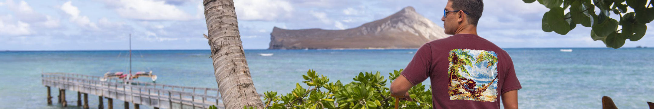 A man wearing a maroon t-shirt with a colorful tropical graphic print on the back, standing on a beach with the ocean, a pier, and a distant rock formation in the background.