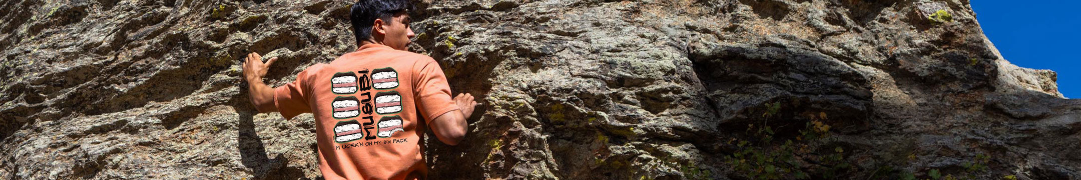 A man in an orange t-shirt with six large spam musubis on the back, climbing a rocky cliff under a blue sky.