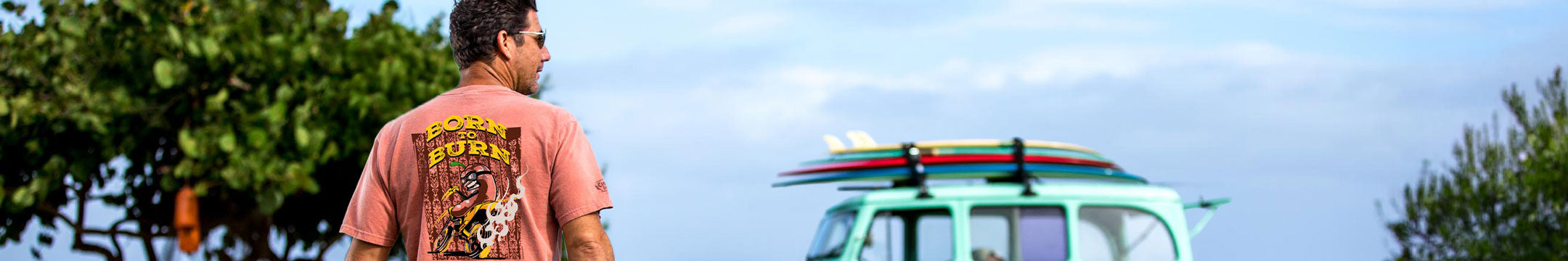 A man wearing a light orange graphic t-shirt stands, looking toward a light blue van with surfboards on top, set against a background of trees and sky.