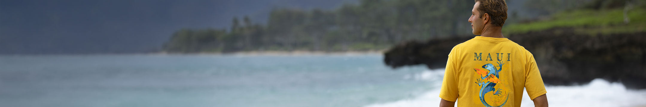 A man wearing a yellow t-shirt with a blue and green graphic and the word 'MAUI' printed on the back, standing on a rocky beach with ocean waves and green cliffs in the background.