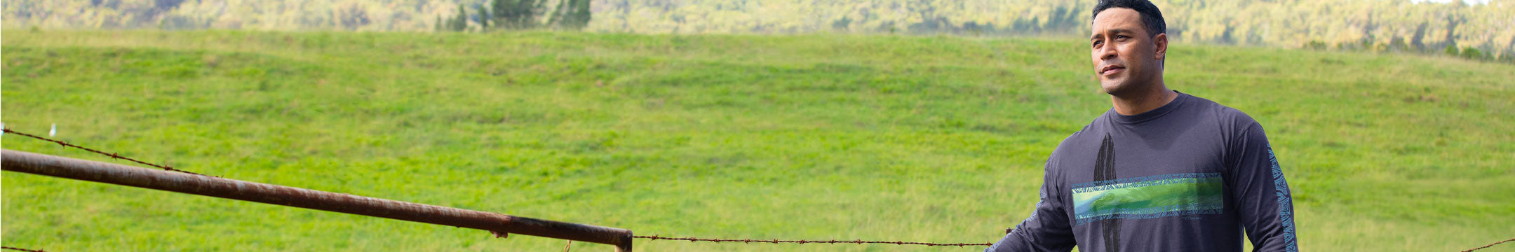 A man wearing a dark gray long-sleeve shirt with a green chest stripe stands in a grassy field next to a wooden fence.