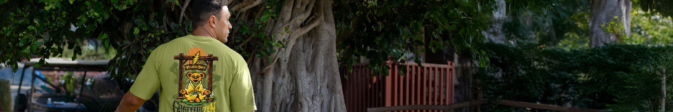 A man viewed from behind wearing a light green t-shirt with a large graphic of a bear with 'GRATEFUL ZEN' print on the back, standing outdoors in front of a large tree trunk and green foliage.