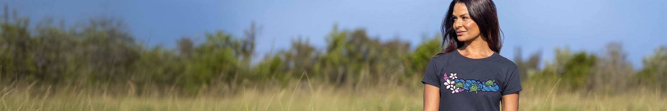 A woman wearing a dark gray t-shirt with a colorful graphic print stands in an outdoor grassy field under a blue sky.