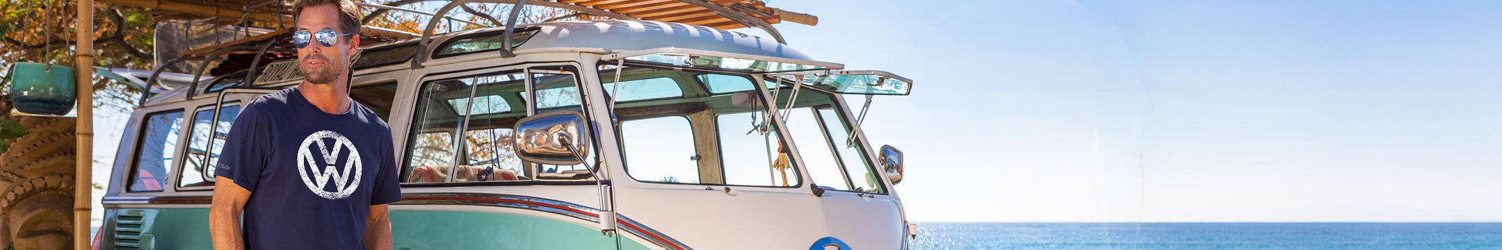 A man wearing a navy blue t-shirt with a white Volkswagen logo stands in front of a vintage cream and teal VW camper van on a beach with the ocean in the background.
