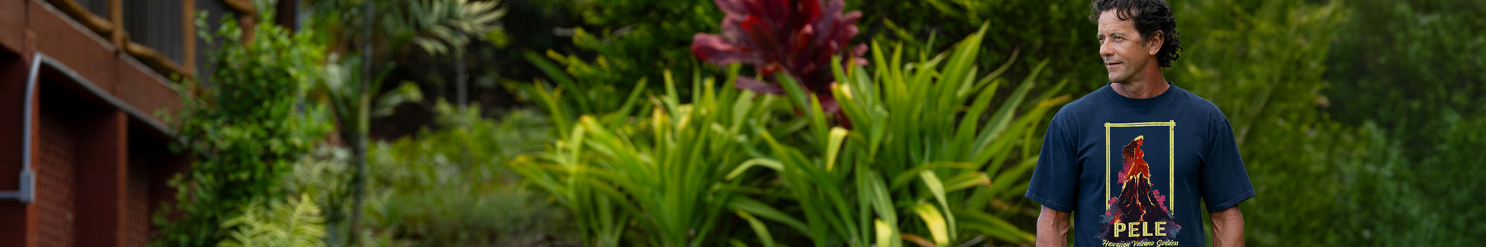 A man wearing a dark navy blue graphic t-shirt with a red and yellow volcano design, standing in a lush tropical garden setting