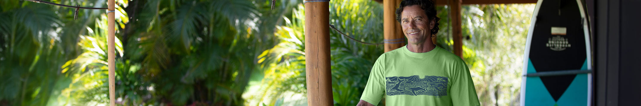 A man wearing a light green t-shirt with a dark graphic print stands in a tropical outdoor setting with palm trees and wooden pillars, with a surfboard visible on the right.