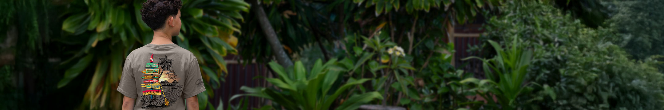 A boy viewed from behind wearing a gray t-shirt with a colorful graphic print, standing in a lush green tropical garden.