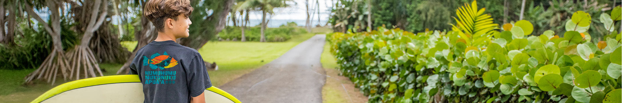 A man wearing a black t-shirt with a colorful graphic fish on the back stands on a paved path in a lush tropical garden, holding a surfboard.