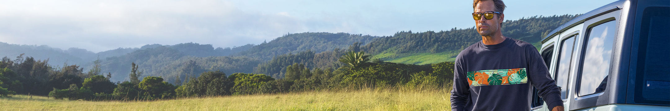 A man wearing a dark gray long-sleeve t-shirt with a colorful graphic print on the front, standing next to a blue vehicle in an outdoor mountain landscape.