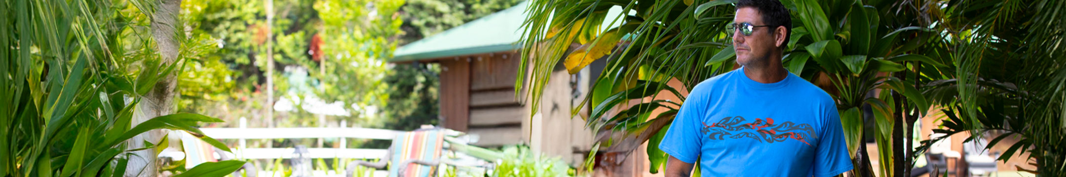 A man wearing a bright blue graphic t-shirt and sunglasses, standing outdoors in a lush green environment with tropical plants and a wooden cabin in the background.